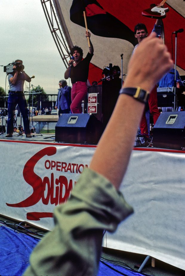 A stage with performing musicians being filmed by a cameramanis skirted with the Operation Solidarity logo. In the foreground a person’s raised arm and clenched fist is seen.