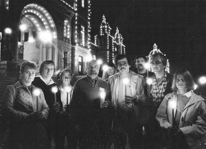 A photograph of nine people holding lit candles face the camera with the BC legislature in the background.
