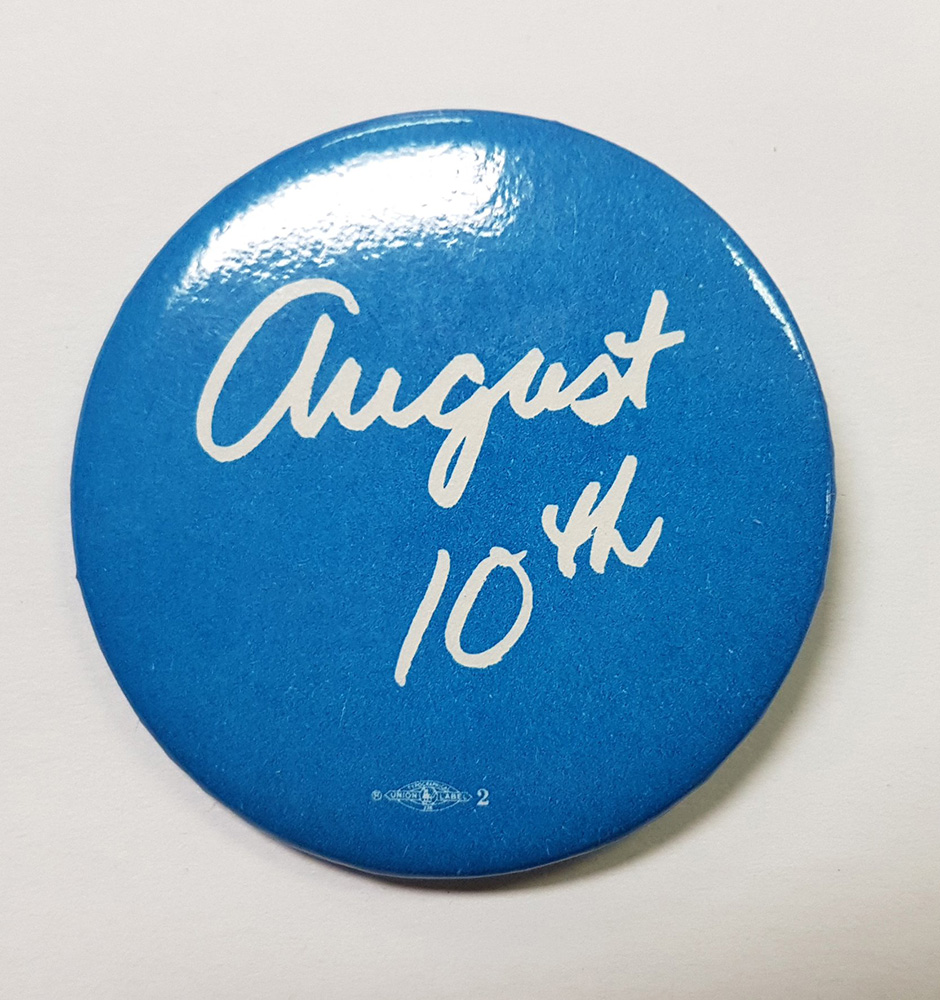 A round blue and white button features script reading “August 10th”. Below the printing can be seen a small union logo.