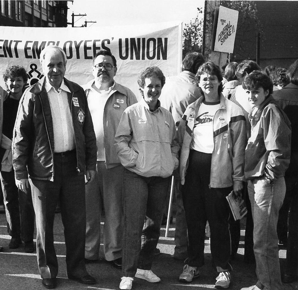 Photograph shows five people wearing Solidarity buttons and shirts pose in front of a banner which reads BC Government Employees’ Union. A sign reading “General Strike!!” can be seen in the background.