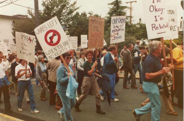 A group of people are marching in a parade. Many hold signs that include “What’s left when you have no rights?”, “Hard Times Won’t Stop us” and “1984 One Year Early!!”