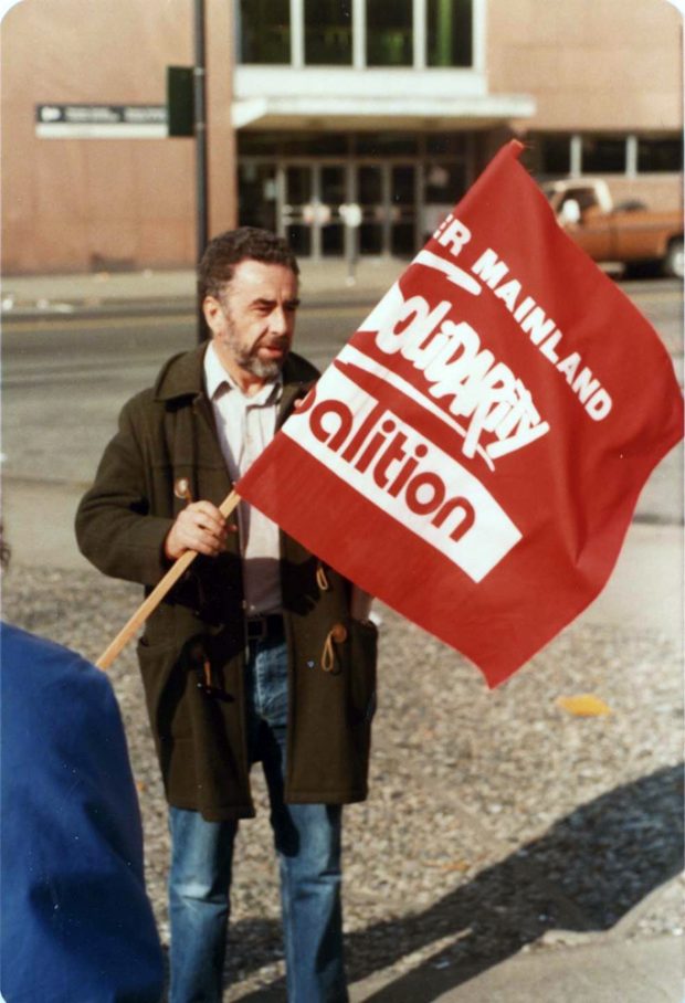 A man holds a partially unfurled red and white flag which reads Lower Mainland Solidarity Coalition.