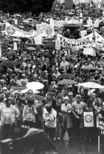 Une foule de personnes font face à la caméra, certains tiennent des parapluies et d’autres des pancartes. On peut lire sur les bannières « Unemployed Teachers Action Centre » (Centre d’aide des enseignants au chômage) et « British Columbia Teachers’ Federation » (Fédération des enseignants de la Colombie-Britannique). 
