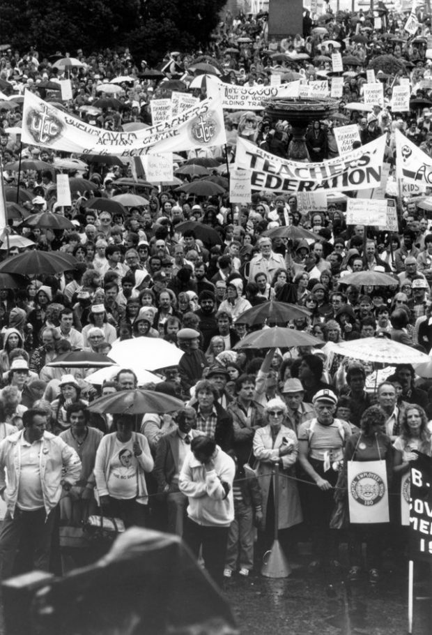 A crowd of people face the camera, some holding umbrellas and others wearing picket signs. Banners read “Unemployed Teachers Action Centre” and “British Columbia Teachers’ Federation”.