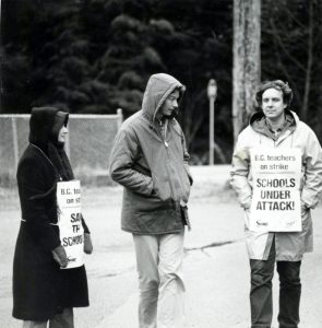 Trois personnes en manteau d’hiver et portant des mitaines marchent le long d’un trottoir. Deux d’entre eux tiennent des pancartes sur lesquelles est inscrit « Les enseignants de la C.-B. en grève, les écoles attaquées! ». Les signes portent les logos de la Solidarity Coalition et de la FECB. 