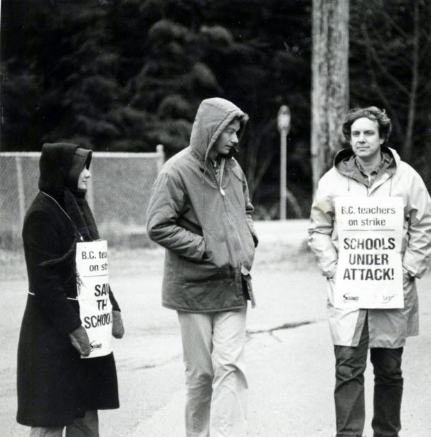 Three people in winter coats and mittens walk along a sidewalk. Two are wearing picket signs reading “BC teachers on strike, Schools Under Attack!”. The signs bear the logos of the Solidarity Coalition and the BCTF.