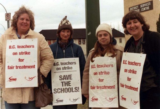 Four women in winter coats and hats stand in a row. They are wearing picket signs reading “BC teachers on strike for fair treatment” and “BC teachers on strike, Save the Schools!”. A sign in the background reads “District Services Centre”.