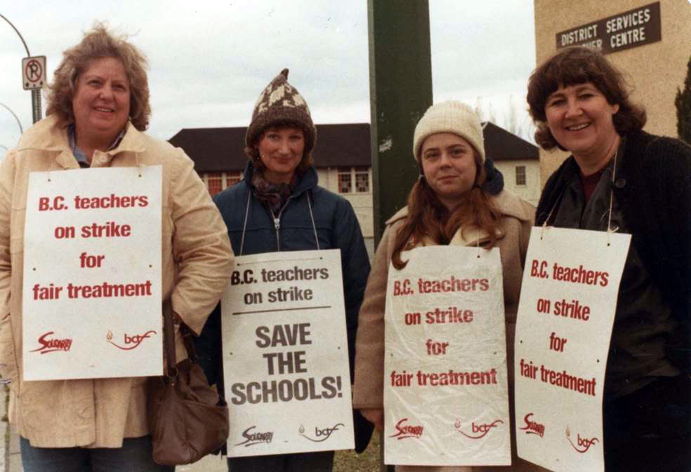 Four women in winter coats and hats stand in a row. They are wearing picket signs reading “BC teachers on strike for fair treatment” and “BC teachers on strike, Save the Schools!”. A sign in the background reads “District Services Centre”.