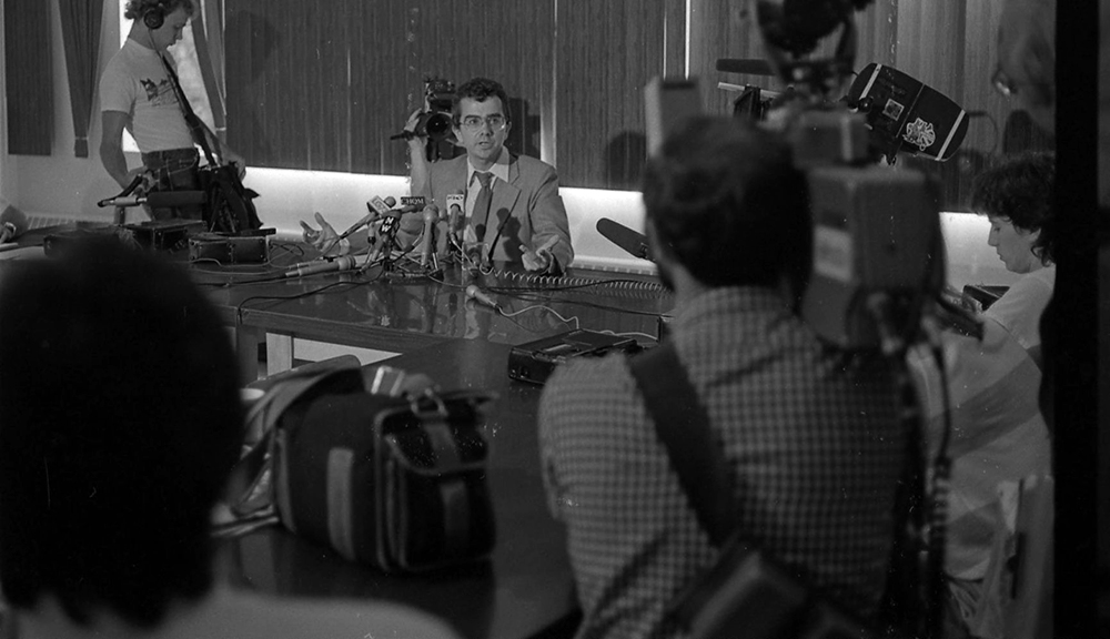 A man is seated at a table with many microphones in front of him. Several television cameras are pointed at him.
