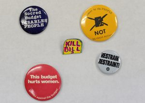 Five round metal buttons, with slogans including: “Restrain restraint!” “The Socred Budget Disables People” and “Government by the PEOPLE NOT the Fraser Institute”
