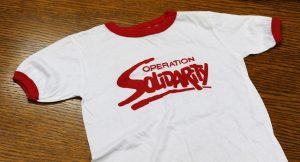 The front of a white t-shirt with a red collar and red Operation Solidarity logo screened on the fabric.