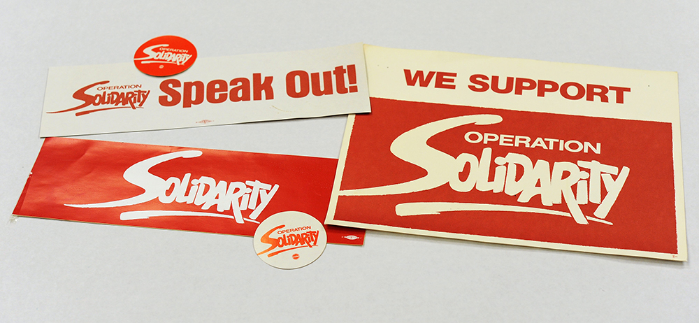 Red and white stickers with the Operation Solidarity logo are displayed. Some include the phrase “Speak Out” and “We Support”.