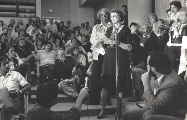 A woman is holding papers and speaking at a microphone. A man stands behind her waiting his turn to speak. They aresurrounded by people on seats and sitting on the floor.