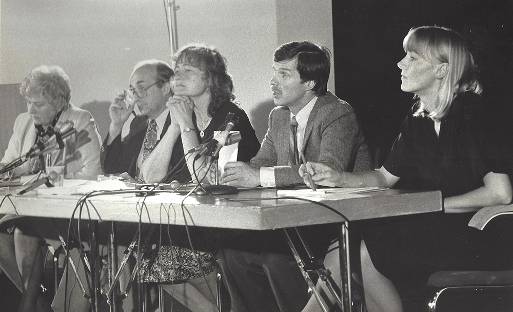 Three women and two men are seated at a table with several microphones in front of them. They appear to be listening to a speaker.