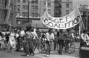 A parade of people is assembled walking, on bicycles and in wheelchairs. A banner reads “Budget Coalition” and other signs are visible.