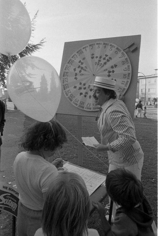 A photograph shows a group of children watching a man spin a carnivalwheel. The children are holding balloons and a sign reads We Support Operation Solidarity.