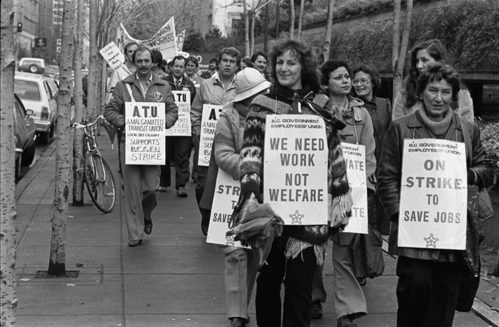 A group of people with picket signs walk along a sidewalk. Signs at the front read “BC Government Employees’ Union, We Need Work not Welfare” and “On Strike to Save Jobs”. Behind them are more people; one sign reads “ATU –Amalgamated Transit Union Local 583 Calgary Supports BCGEU strike”.