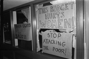 Deux fenêtres avec des affiches manuscrites indiquent « Les droits de la personne ne sont pas à vendre! », « Ce budget nuit aux femmes! » et « Cessez les attaques contre les pauvres! » 