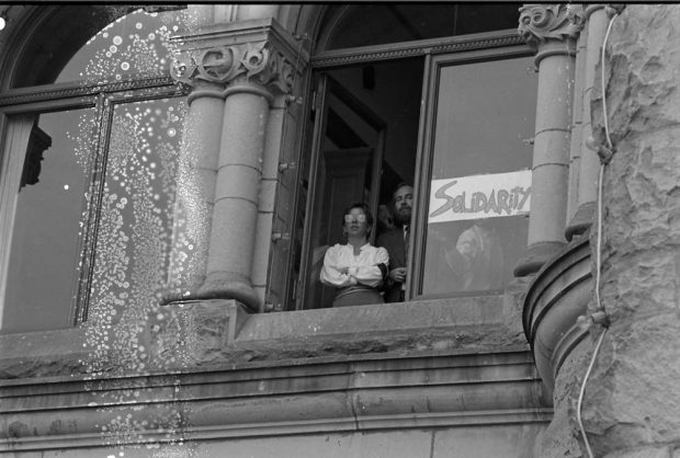 Two people are pictured at an open window and a hand lettered sign reads “Solidarity”.