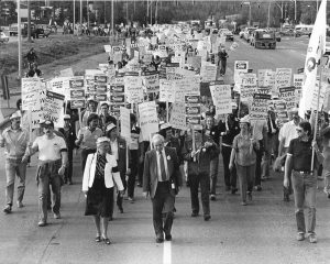 Un grand groupe de personnes paradent sur une rue pavée en tenant des pancartes lisant « En soutien à Operation Solidarity », « Bill où est passé ton sens de la valeur » et « Grace McCarthy maltraite les enfants ».