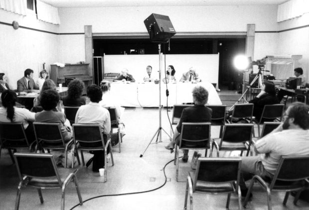 Four people are seated at a table facing a seated audience. A camera, lights and audio equipment are seen.