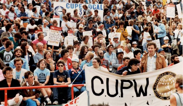 Stadium seats are filled with people and banners from Canadian Union of Postal Workers and Canadian Union of Public Employees are visible.