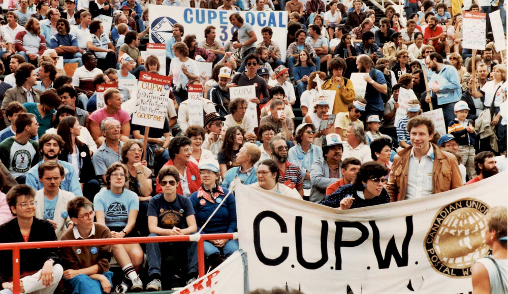 Stadium seats are filled with people and banners from Canadian Union of Postal Workers and Canadian Union of Public Employees are visible.