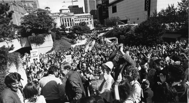 A large group of people stand and cheer outside a building holding signs reading “United We Stand” and “People Not Power”. The Vancouver Art Gallery can be seen in the background.