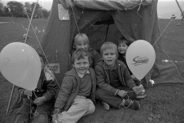 Five young children sit on the grass at the entrance to a small tent. They are holding balloons with the Operation Solidarity logo printed on them.