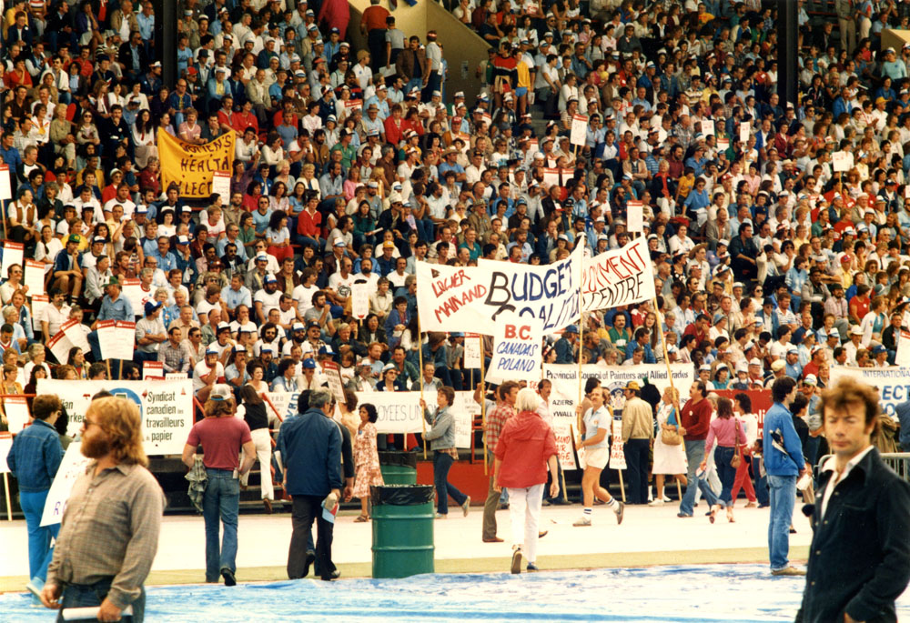 A photograph shows a crowd of people seated in a stadium. Several people parade past carrying banners that read “Lower Mainland Budget Coalition” and “BC Canada’s Poland”