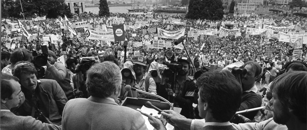 A man, speaking at a podium, faces out towards a large crowd of people on the lawn of the B.C. legislature. Reporters are gathered, holding microphones and cameras towards the podium. Organizational banners and protest signs are visible throughout the crowd.