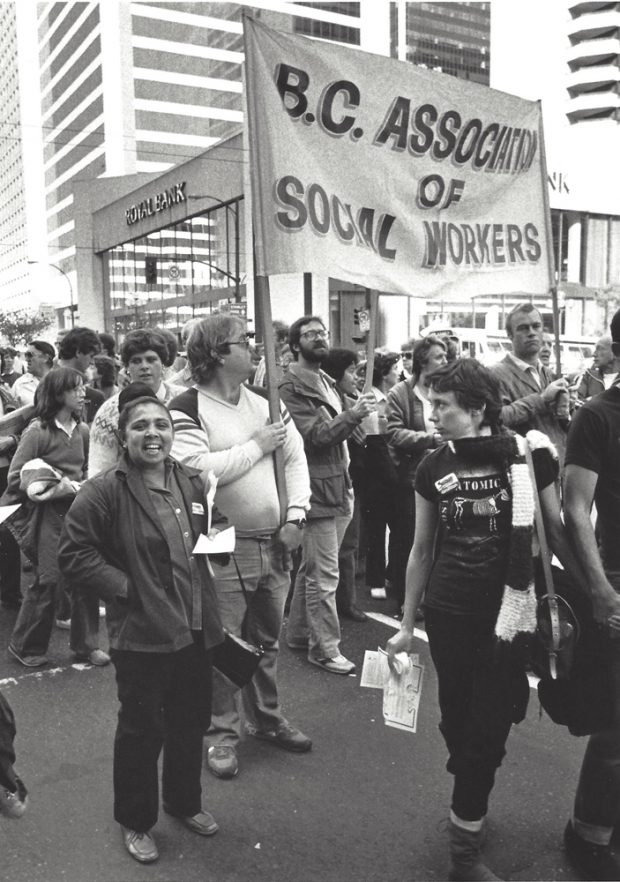 Two men hold a banner reading “B.C. Association of Social Workers”. They are part of a parade of people standing on a street. Some are wearing Solidarity Coalition buttons.