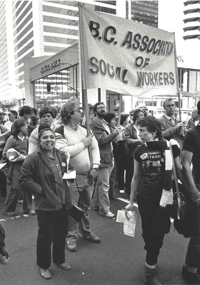 Deux hommes tiennent une bannière intitulée « B.C. Association of Social Workers » (Association des travailleurs sociaux de la C.-B.).  Ils font partie d’un défilé de personnes se tenant debout dans la rue. Certains portent des macarons de la Solidarity Coalition. 