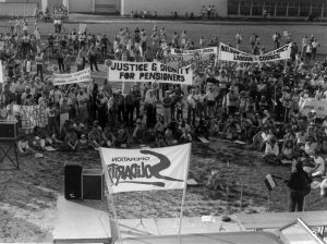 A stage faces a group of people standing and sitting on the grass. The Operation Solidarity flag flies and banners include “Justice & Dignity for Pensioners”, “Nelson Womens Centre” and “Nelson and District Labour Council”