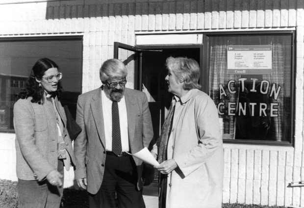 Three people in a conversation stand outside an office storefront. The window of the office displays a federal government sign and the words Action Centre.