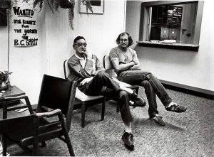 Deux hommes sont assis les jambes et bras croisés dans une des salles d’attente d’un bâtiment. Une affiche sur le mur lit « Bill Bennett, recherché pour le meurtre de l’Esprit de la C.-B. ». 
