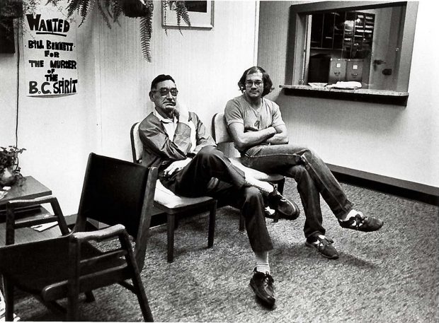 Two men are seated with their legs and arms crossed in a reception area of a building. A sign on the wall reads “Wanted Bill Bennett for the Murder of the BC Spirit”.