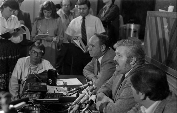 Three men sit at a table with many microphones in front of them. Reporters with notebooks are in the background.