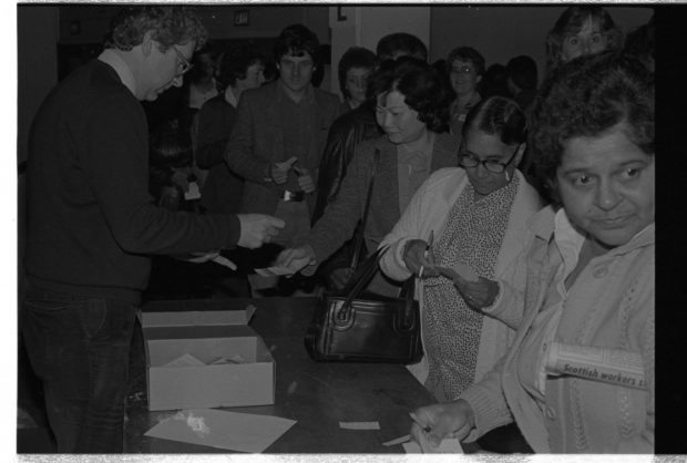 A man stands behind a table with a box. Women and men line up to in front of the table holding papers and pens.
