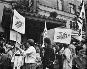 De nombreuses personnes défilent devant l’entrée principale de l’Hôtel Vancouver. Ces personnes tiennent des pancartes sur lesquelles on peut lire « Préparez la grève générale!! » et portent un drapeau d’Operation Solidarity. 
