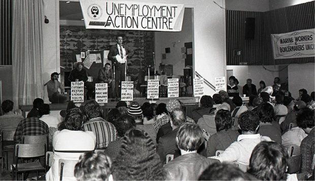 A man stands at a microphone to address an audience from a stage. Three men are seated on the stage . A banner above the stage reads “Unemployment Action Centre” and other signs are taped to the stage.