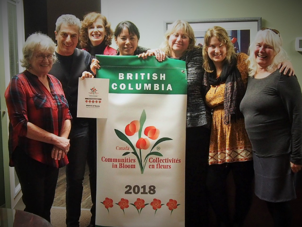 A group of seven people standing around a vinyl banner, with woman at centre holding it up. The banner reads: British Columbia Canada Communities in Bloom Collectivitiés de Fleurs 2018 with a floral design.
