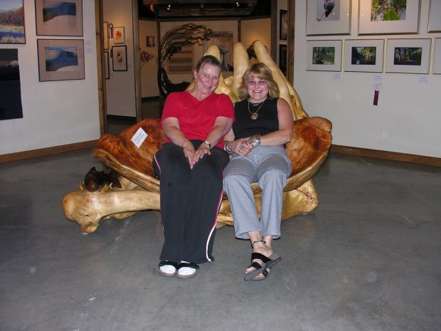 Two women smiling and sitting on a large love seat-shaped sculpture made of wood.