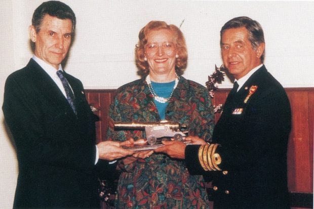 Two men in suits on either side of a woman, presenting her with a ship's cannon.