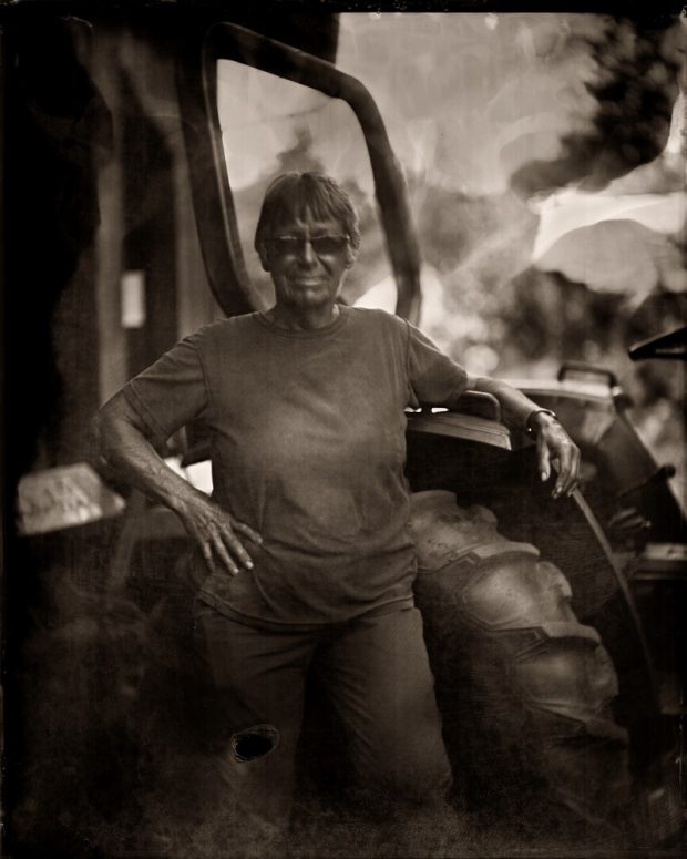 A sepia photograph of a woman standing beside a tractor with her arm resting on the open door.