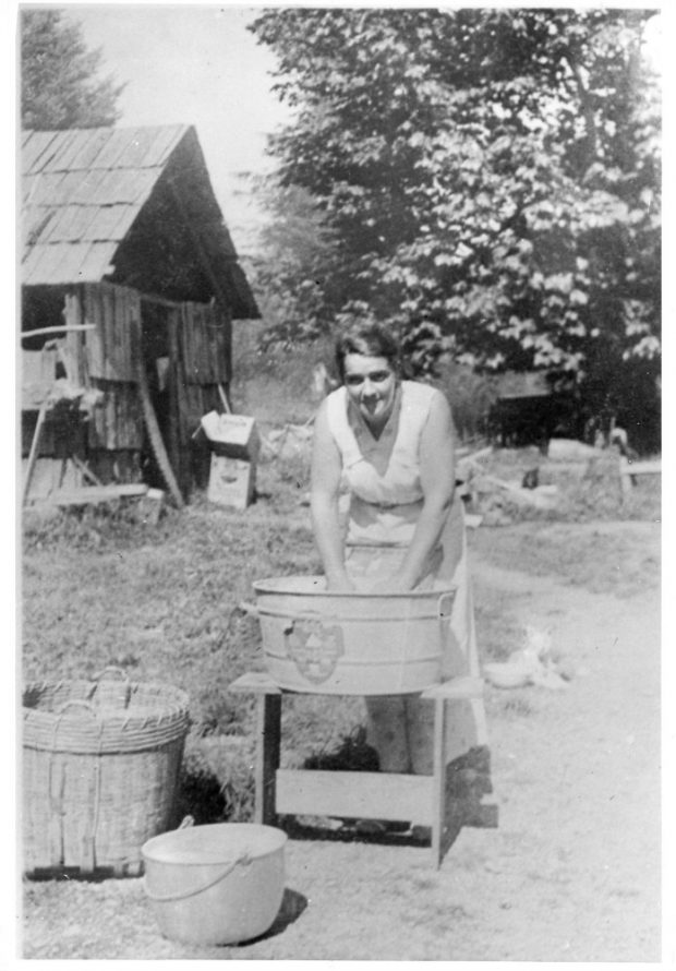 A black and white photograph of a woman washing something in a large metal tub with a washboard.