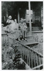 Photographie en noir et blanc d’une femme âgée dans une chaise berceuse en haut des marches d’une véranda; une femme et son mari tenant un bébé sont assis un peu plus bas dans les marches.