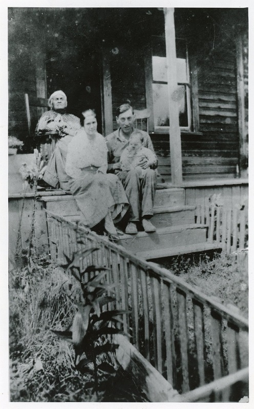 A black and white photograph of a woman in rocking chair at the top of verandah steps with a woman and husband holding a baby sitting on the steps below.