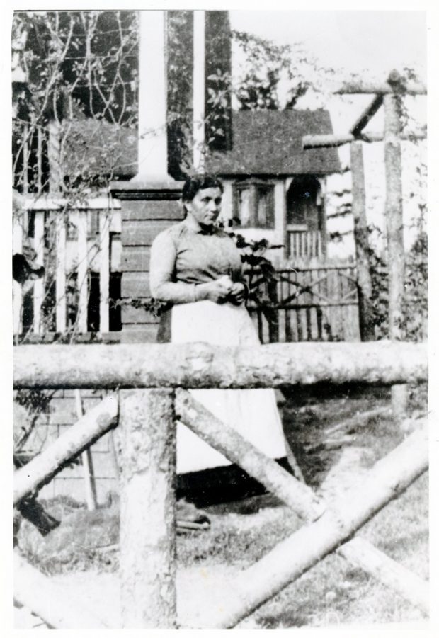 A black and white photograph of a woman standing in a yard between a wooden gate and a house.
