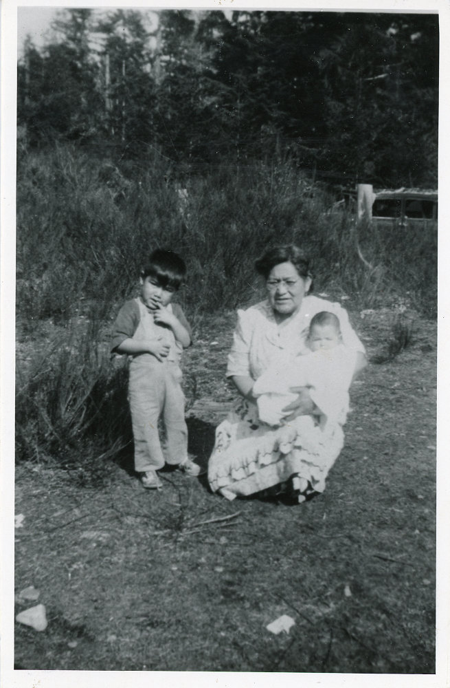 A woman kneeling outdoors holding a baby, with a boy standing beside on her right.